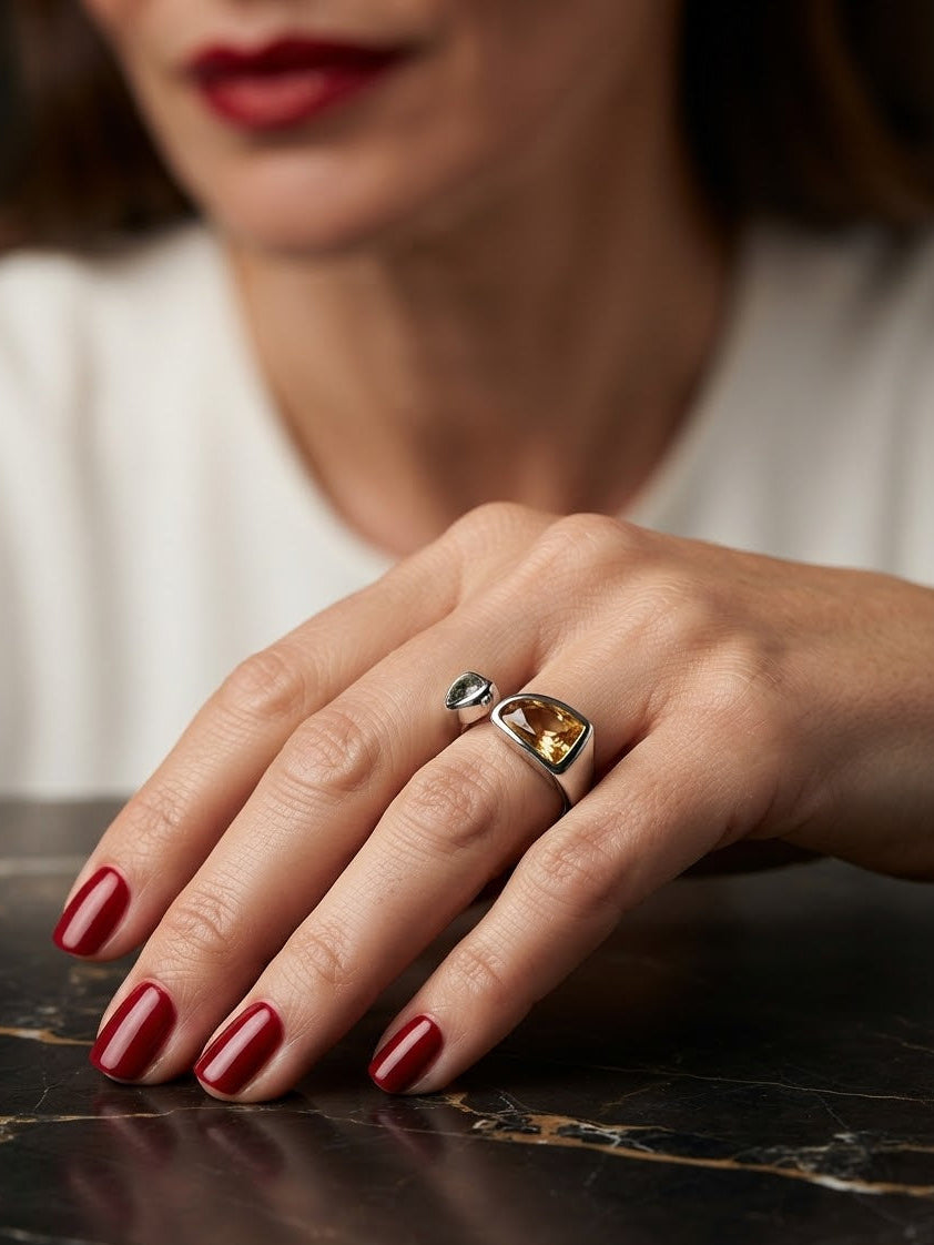 A close-up of a woman's hand resting on a dark marble surface, wearing a silver ring with a square-cut amber gemstone and a small dark stone beside it.