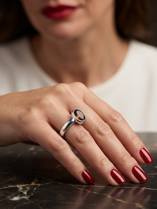 A close-up of a woman's hand resting on a dark marble surface, wearing a silver ring with a round amber-colored stone. The model has red lipstick and red manicured nails, and her white top is softly blurred in the background.