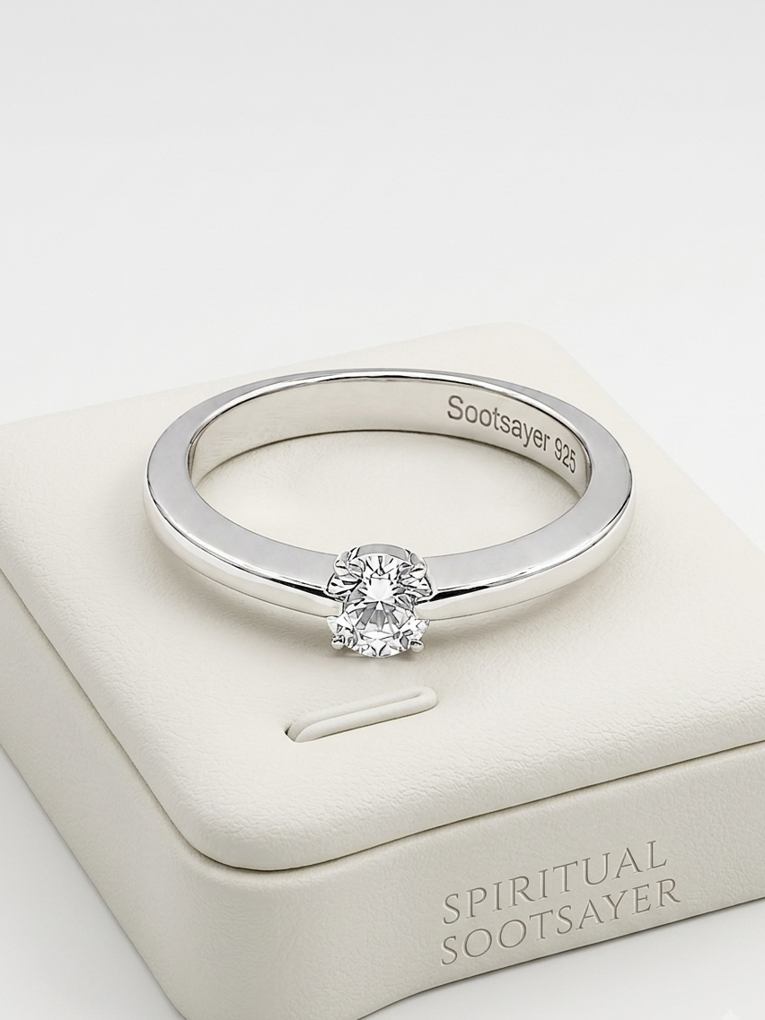A silver solitaire engagement ring with a round-cut clear gemstone, displayed on a cream-colored Spiritual Sootsayer jewelry box. The inside of the band is engraved with 'Sootsayer 925'.