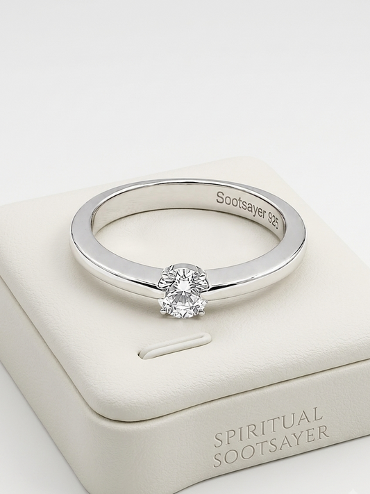 A silver solitaire engagement ring with a round-cut clear gemstone, displayed on a cream-colored Spiritual Sootsayer jewelry box. The inside of the band is engraved with 'Sootsayer 925'.