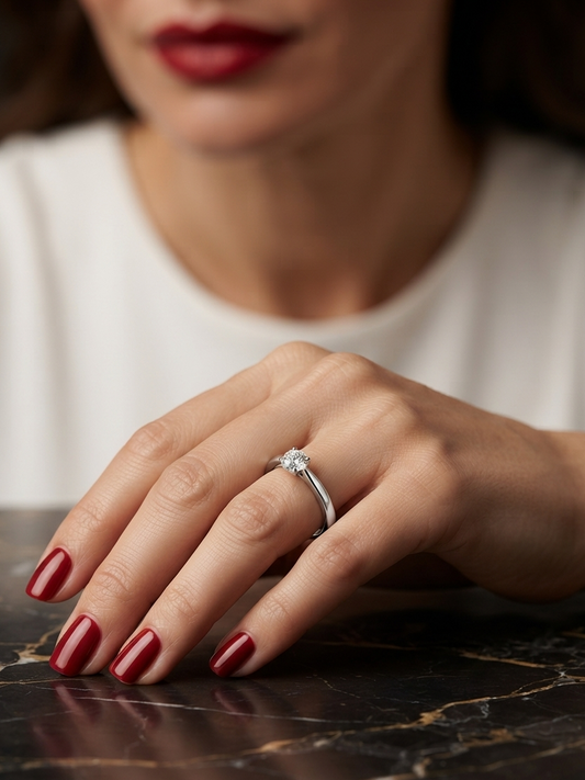 A close-up of a woman's hand resting on a dark marble surface, wearing a silver ring with a clear round gemstone. The background shows a softly blurred woman in a white top with dark red lipstick and nail polish.