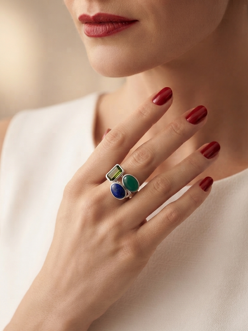 A close-up lifestyle shot of a model wearing a silver ring with three colorful stones (green, blue, and dark blue) on her finger. The model has red nail polish and is wearing a white sleeveless top against a soft, warm beige background.