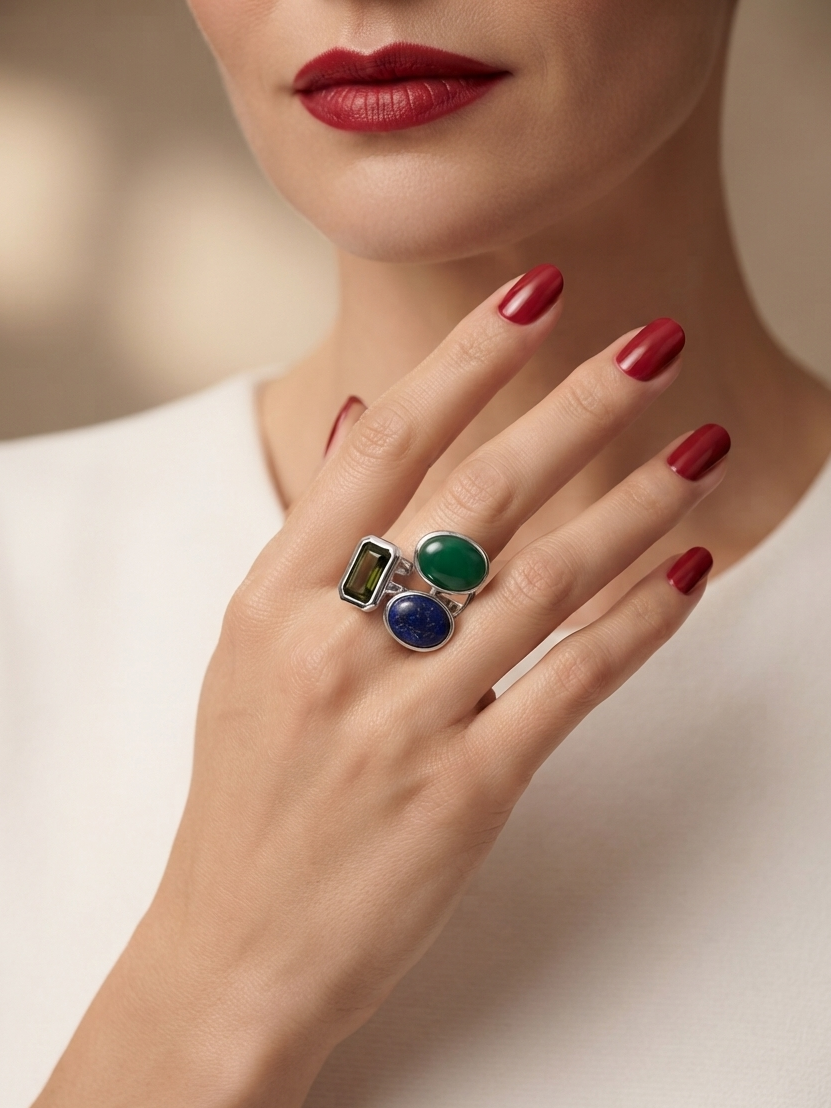 A close-up of a model's hand wearing a silver ring with three colorful stones: a green oval, a blue oval, and a rectangular green stone. The model has red nail polish and is wearing a white top against a soft, warm-toned blurred background.