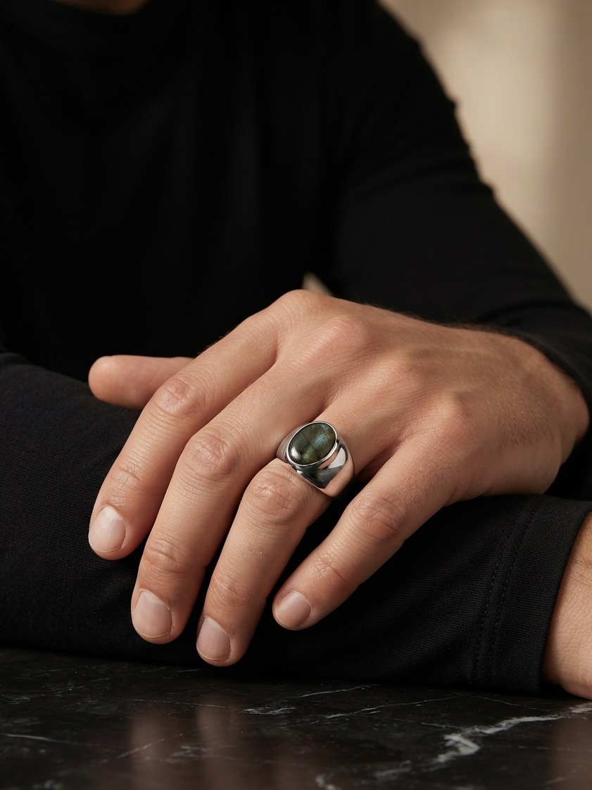 A close-up of a person's hand resting on a dark marble surface, wearing a silver-toned signet ring with a round green stone.
