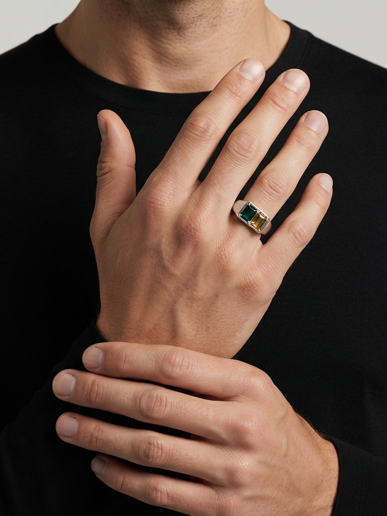 A close-up of a man's hands wearing a gold ring with a rectangular green gemstone, set against a black shirt and a neutral gray background.