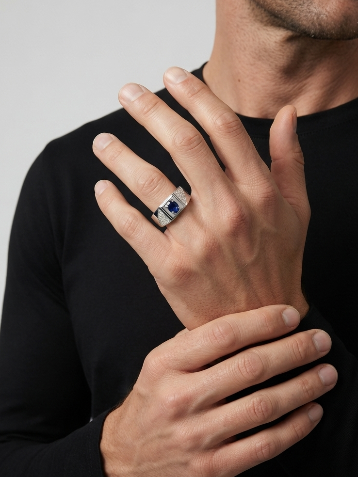 A close-up of a man's hands wearing a silver ring with a rectangular blue gemstone, set against a plain light gray background.