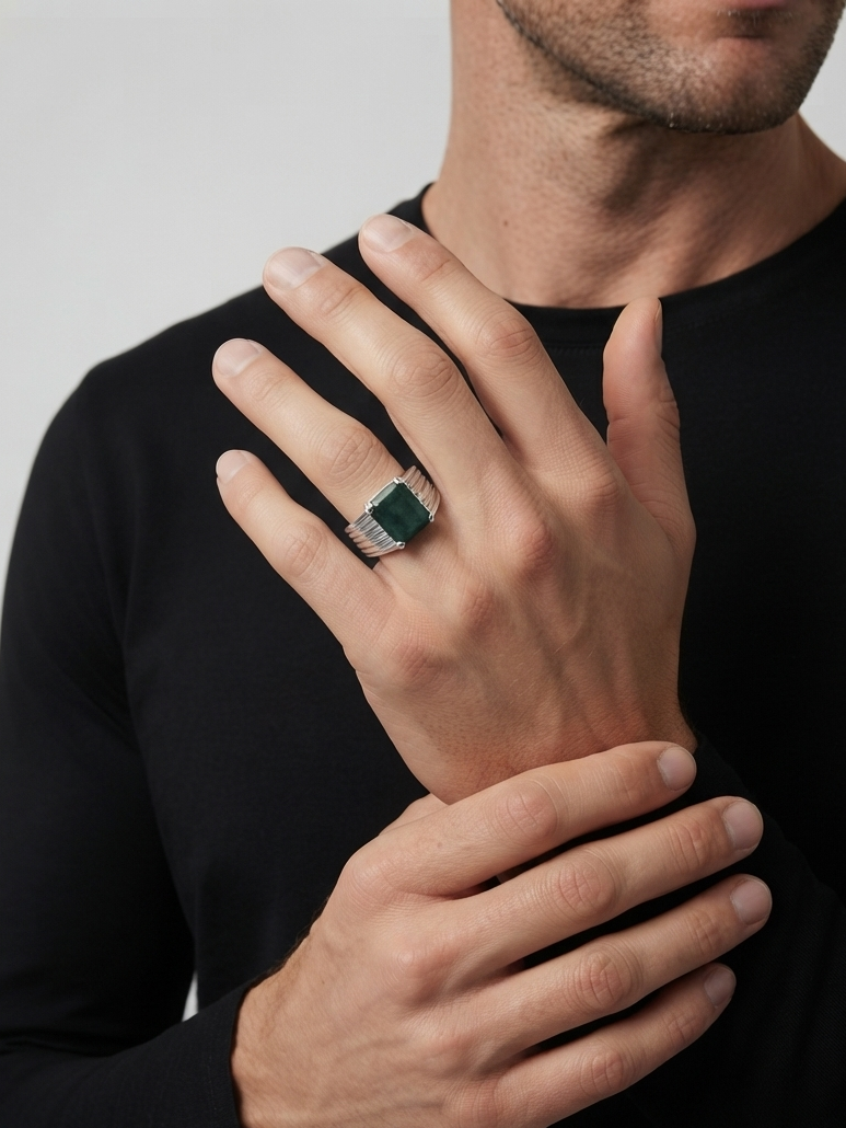 A close-up of a man's hand wearing a silver-toned signet ring with a rectangular dark green stone, set against a black long-sleeve shirt and a light gray background.