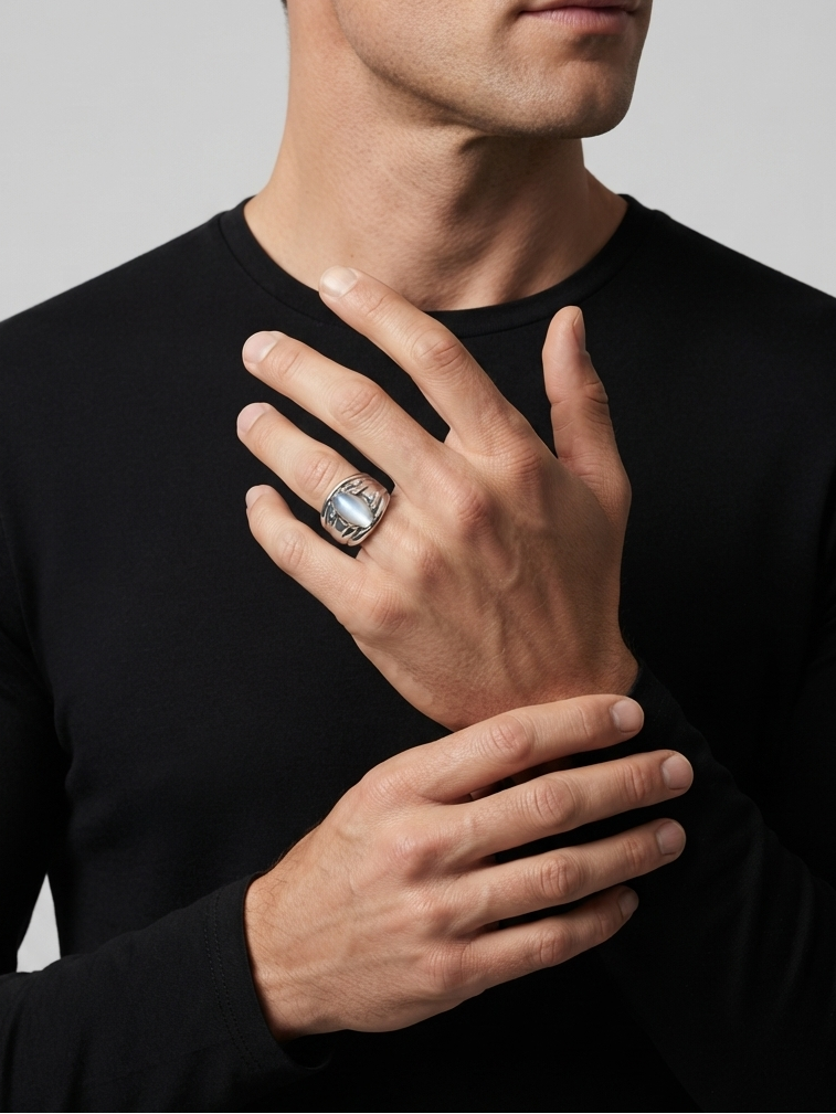 A close-up of a man wearing a black long-sleeve shirt and a large silver-toned ring with a dark stone on his ring finger, set against a plain light gray background.