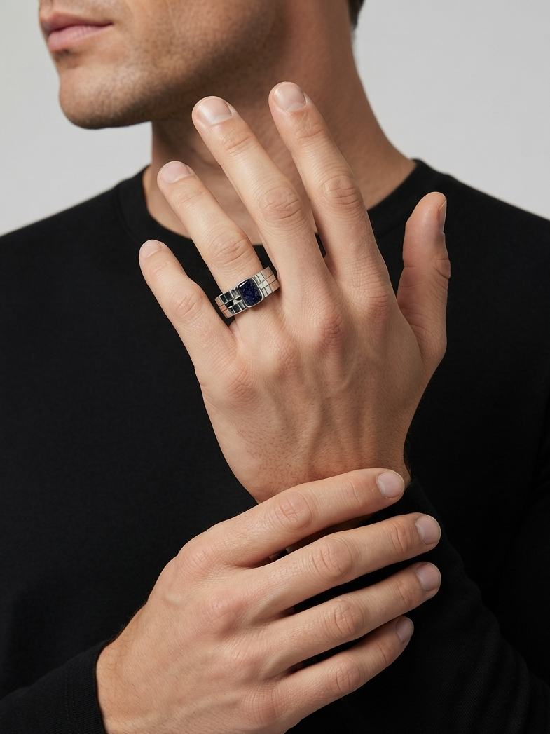 A close-up of a man's hand wearing a silver band ring with a central dark blue rectangular stone, set against a neutral gray background.