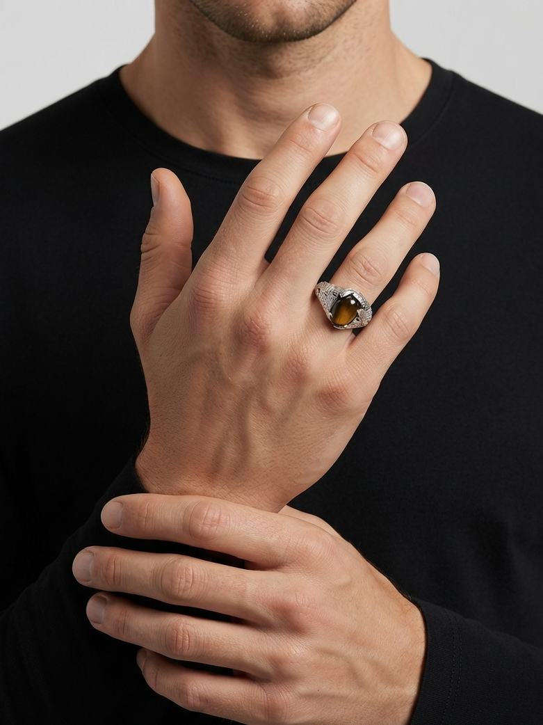 A close-up of a man's hands wearing a silver-toned ring with a large, polished amber-brown stone, set against a black long-sleeve shirt and a light gray background.