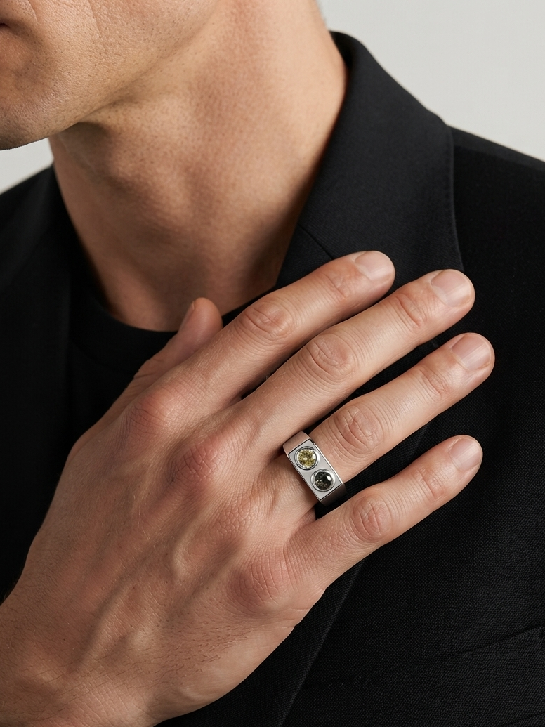 A close-up of a man's hand wearing a silver ring with two small round stones, one gold and one green, styled on his index finger while wearing a black blazer.