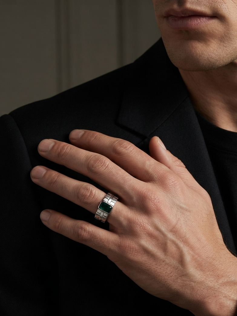 A close-up of a man's hand resting on his black blazer, wearing a silver-toned ring with a rectangular green gemstone.