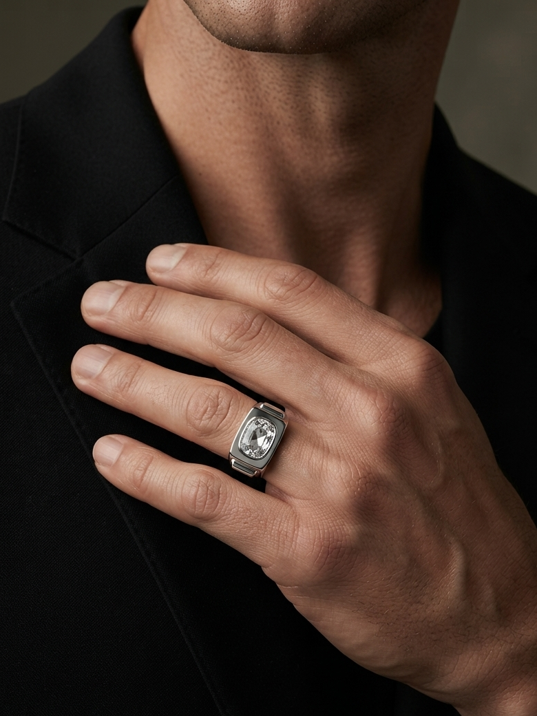 A close-up of a man's hand wearing a silver-toned signet ring with a clear central gemstone, set against a black blazer and a dark gray background.