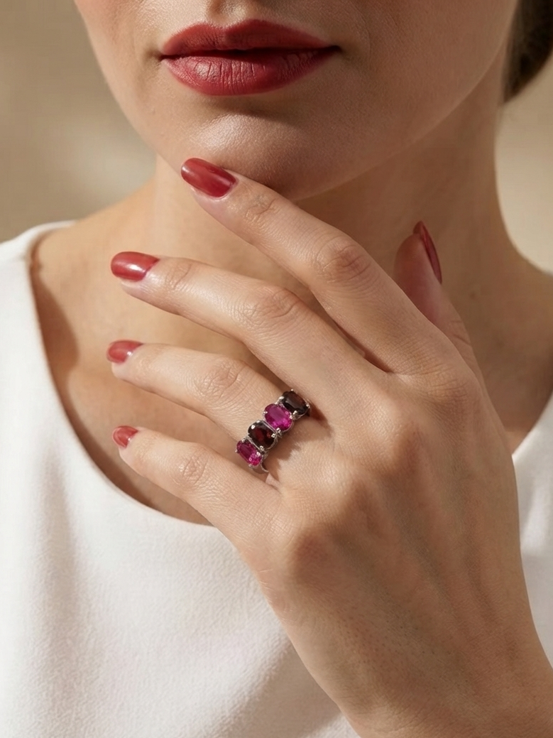 A close-up lifestyle shot of a model wearing a silver ring set with three pink gemstones, resting on her index finger. She is wearing a white top and has red nail polish.