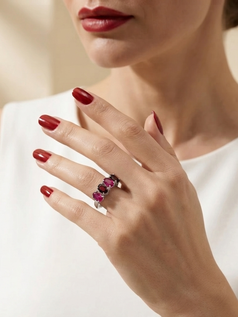 A close-up lifestyle shot of a model wearing a silver ring with three pink gemstones on her finger, paired with a white sleeveless top and dark red nail polish.