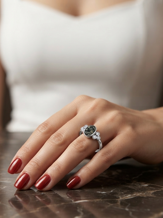 A close-up lifestyle shot of a woman's hand resting on a dark marble surface, wearing a silver ring with a dark green oval gemstone and small accent stones.