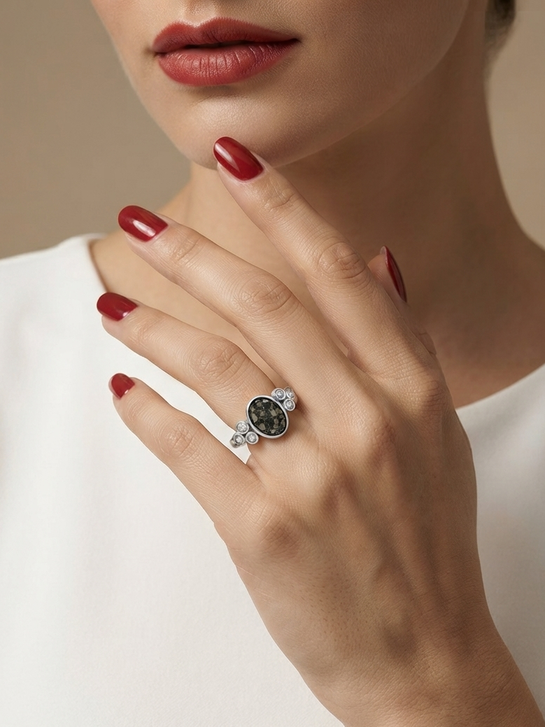 A close-up lifestyle shot of a model's hand wearing a silver ring with a dark green oval stone and small clear accent stones, set against a soft beige background and white top.