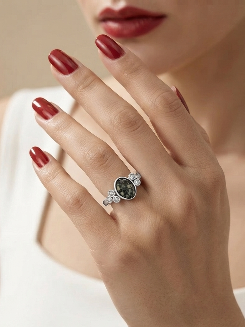 A close-up of a model's hand wearing a silver ring with a dark green oval stone and small clear accent stones, set against a soft beige background.