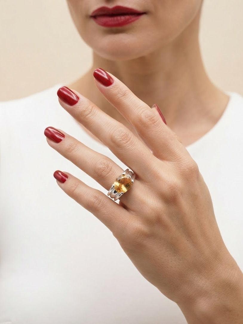 A close-up of a woman's hand wearing a silver ring with a central yellow gemstone, set against a soft beige background and white top.
