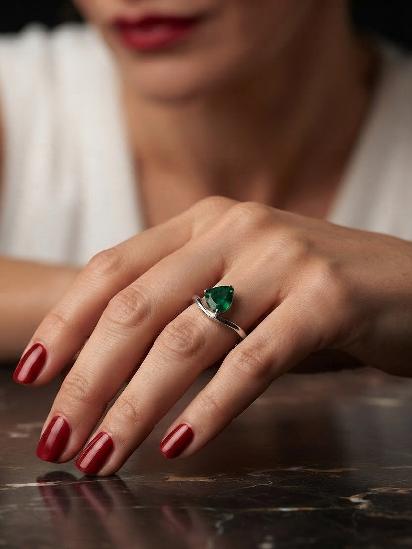 A silver ring with a triangular green gemstone worn on a woman's hand, which has dark red nail polish, resting on a dark marble surface.