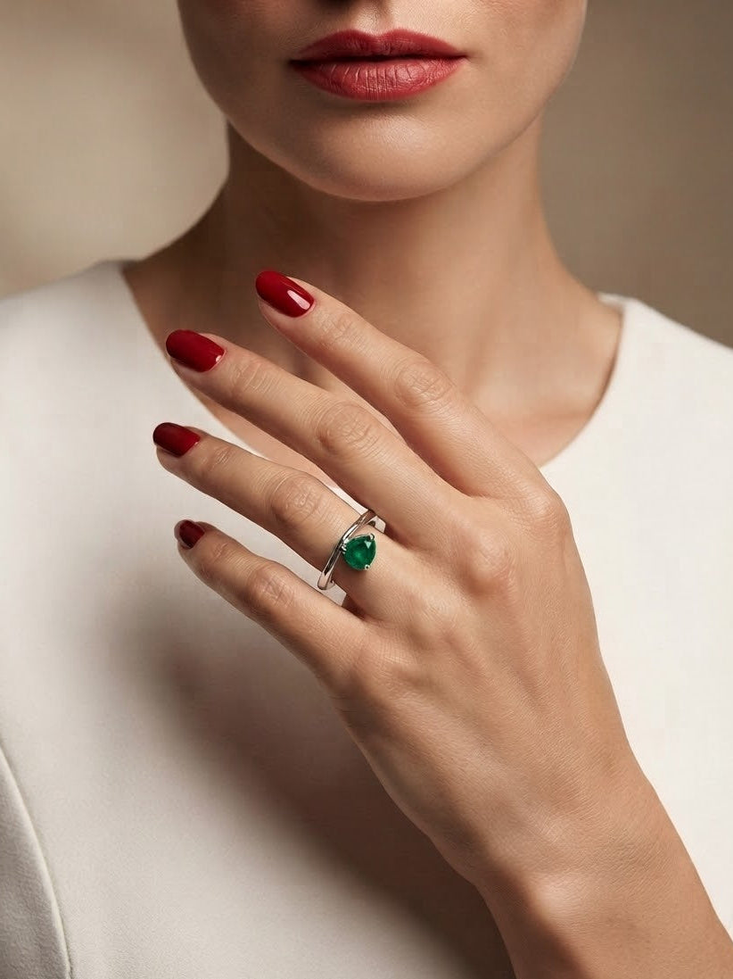 A close-up of a model's hand wearing a silver ring with a marquise-cut green gemstone, set against a soft beige background and white top.