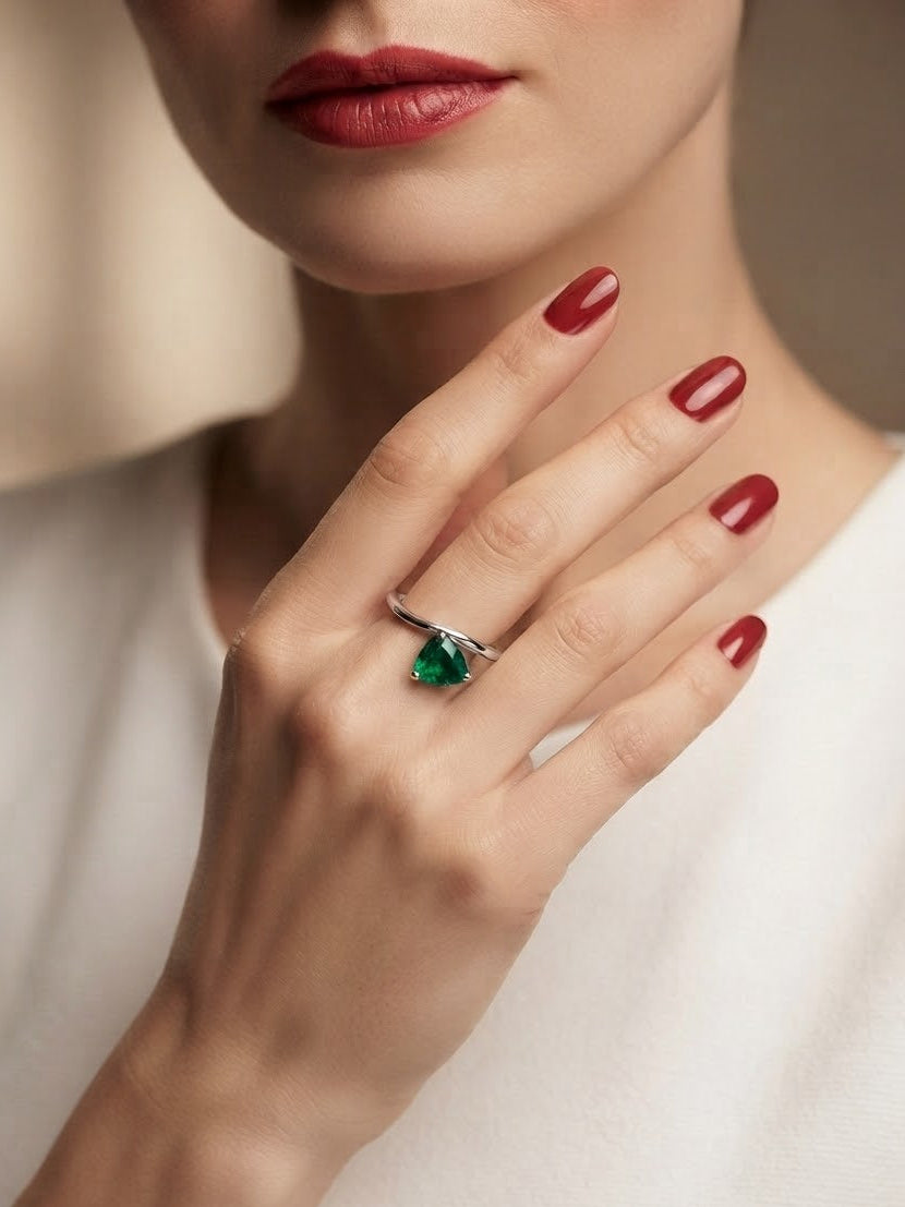 A close-up lifestyle shot of a model's hand wearing a silver ring with a pear-cut green gemstone, set against a soft beige background.