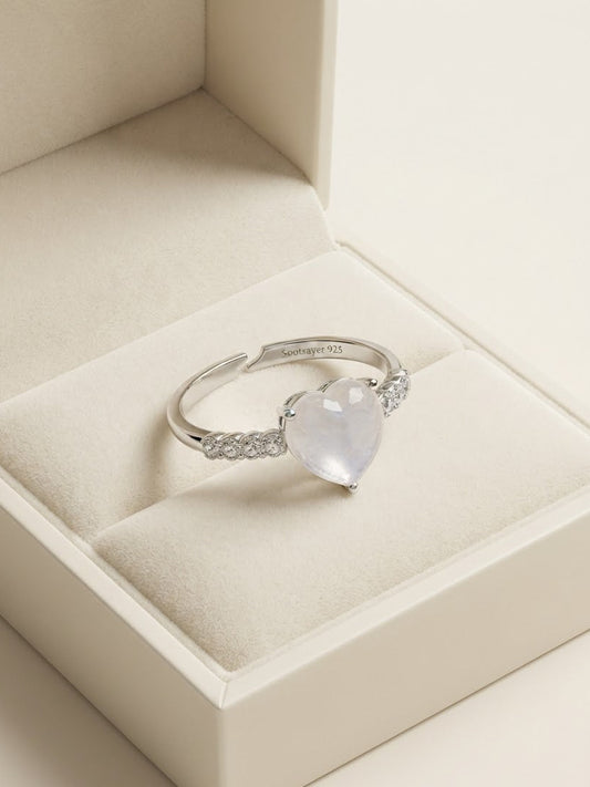 A silver-toned ring with a heart-shaped clear gemstone and small side stones, displayed on a cream-colored jewelry box.