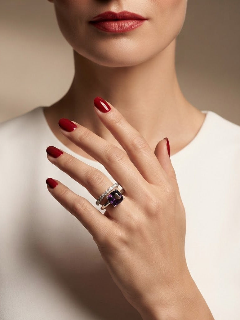 A close-up of a model's hand wearing a silver ring with a dark purple rectangular gemstone, set against a soft beige background and white top.