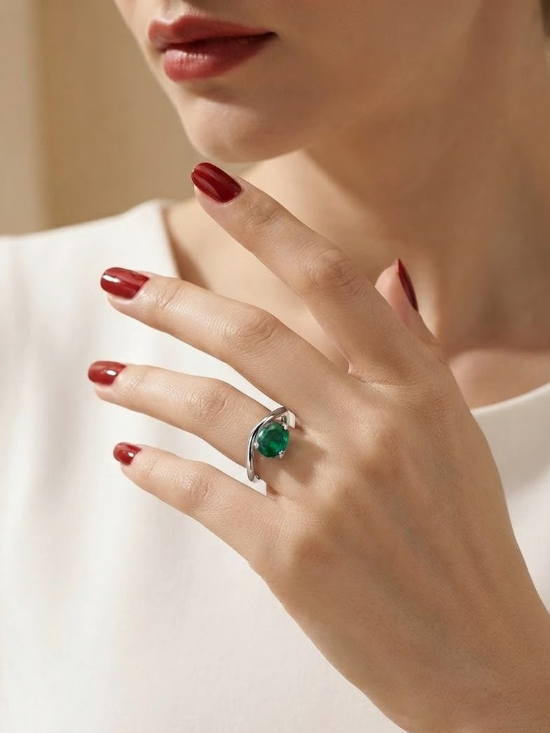 A close-up of a woman's hand wearing a silver ring with a heart-shaped green gemstone, set against a soft beige background and white top.
