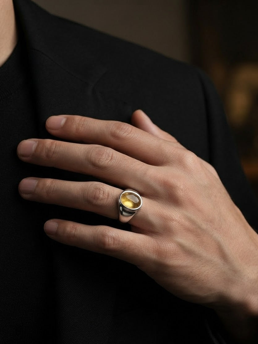 A silver-toned men's ring with a bezel-set round yellow gemstone, shown worn on a man's ring finger against a black shirt.