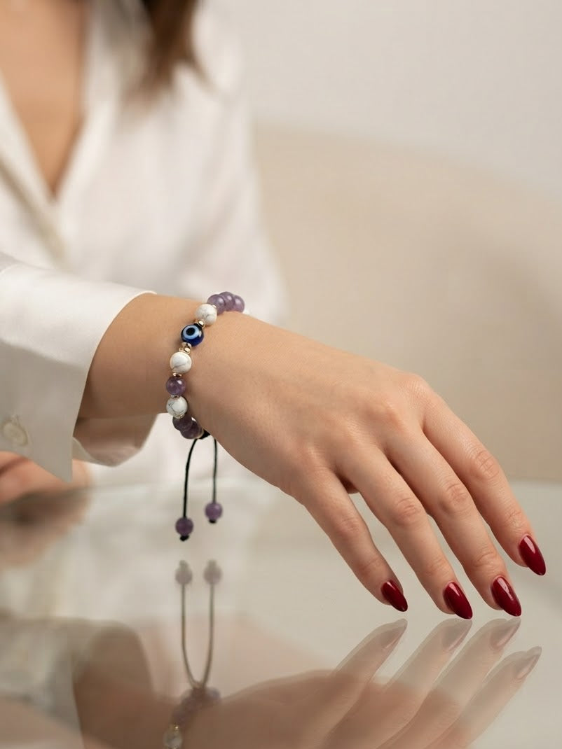 A close-up of a person's hand wearing a beaded bracelet with purple, white, and blue evil eye beads, styled with a white shirt and red manicured nails.