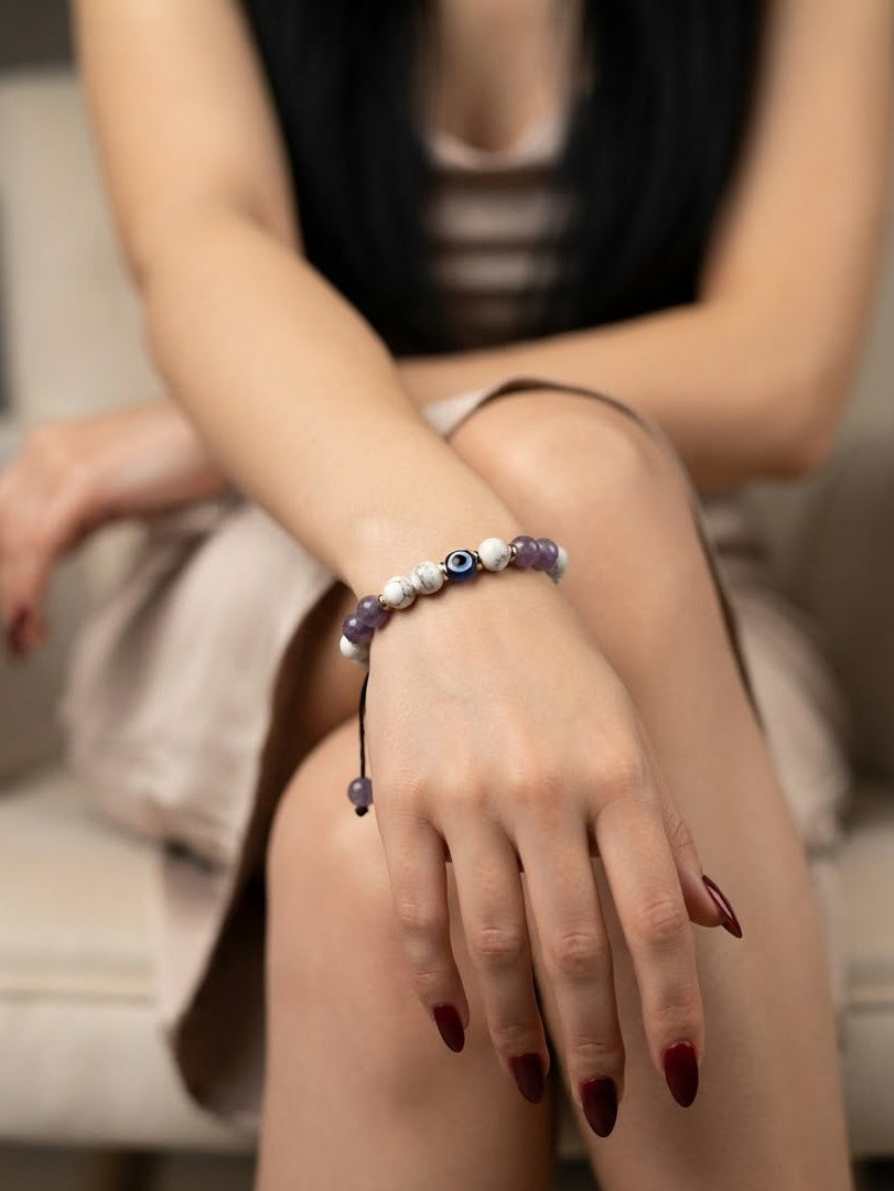 A close-up of a person's hand and wrist wearing a beaded bracelet with white pearl-like beads, purple beads, and a central evil eye motif, seated in a soft indoor setting.