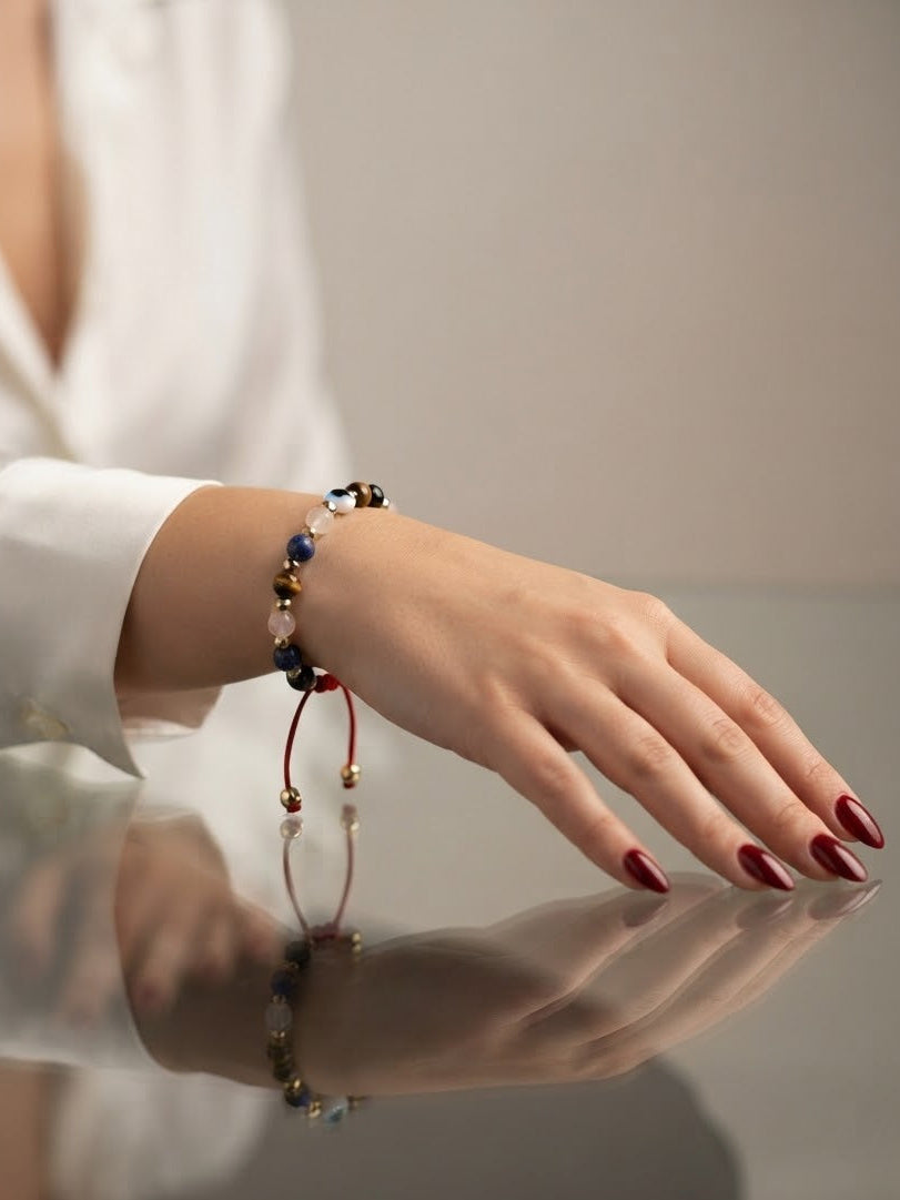A close-up of a person's hand wearing a beaded bracelet with a red cord and dangling bead charm, resting on a reflective surface.