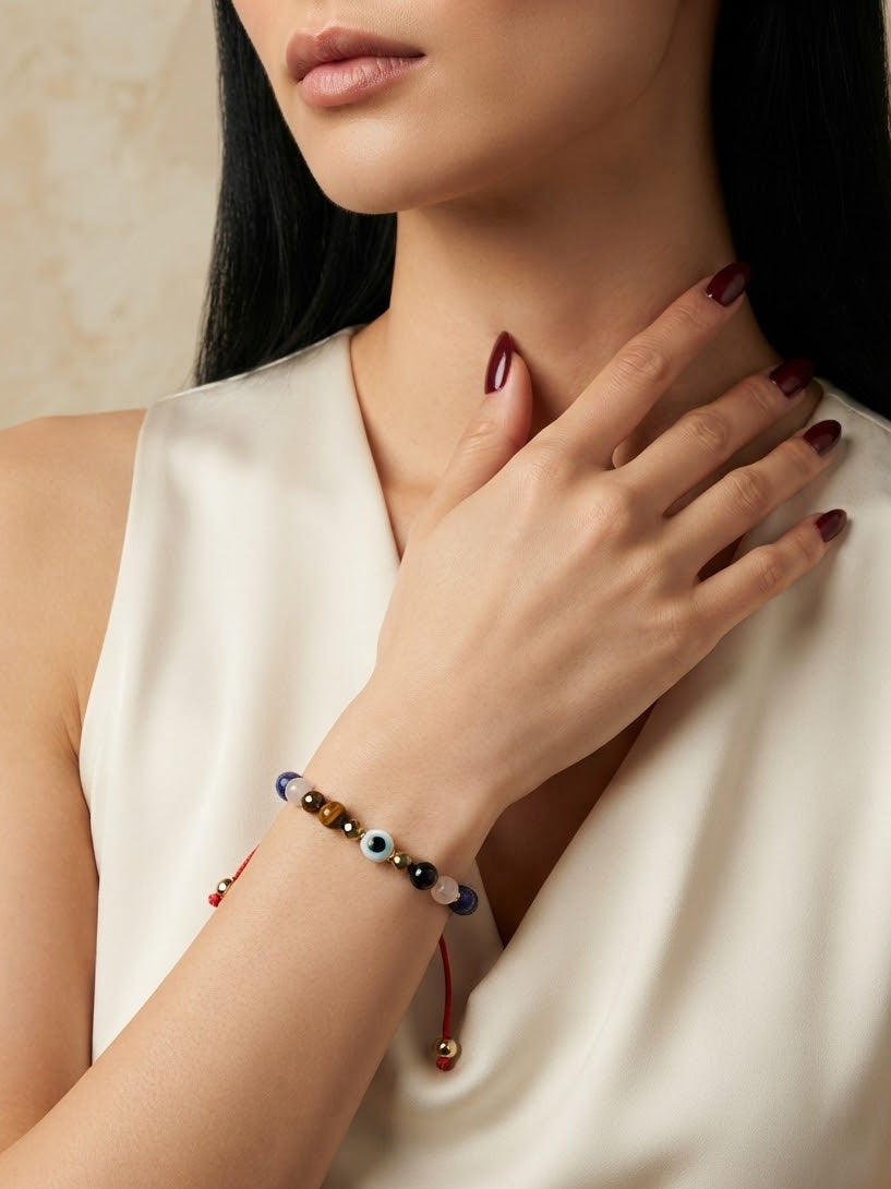 A close-up of a woman wearing a beaded charm bracelet on her wrist, paired with a cream satin top. The bracelet features colorful beads and a red cord with a small gold bead.