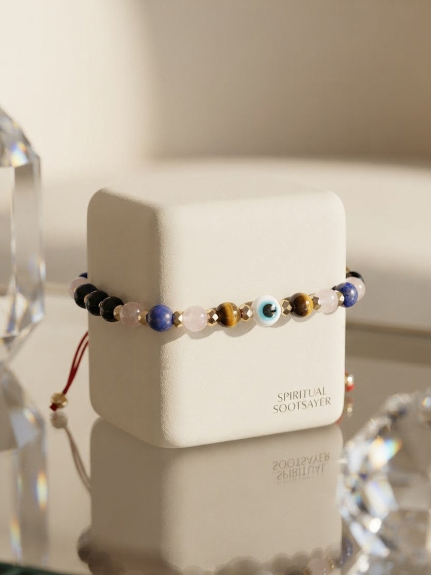 A beaded spiritual socks saver bracelet displayed on a white branded jewelry box, set on a reflective glass surface with crystal props.