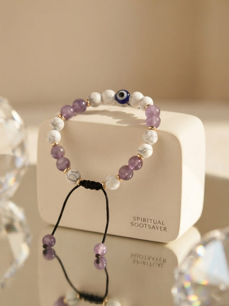 A beaded stretch bracelet with purple amethyst, white marble, and a blue evil eye bead, displayed on a cream-colored box labeled 'SPIRITUAL SOOTSAYER'.