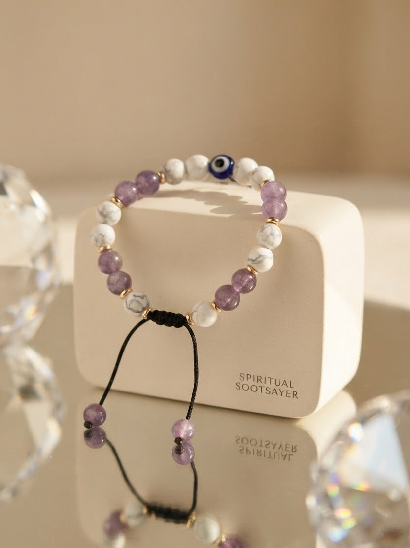 A beaded stretch bracelet with purple amethyst, white marble, and a blue evil eye bead, displayed on a cream-colored box labeled 'SPIRITUAL SOOTSAYER'.