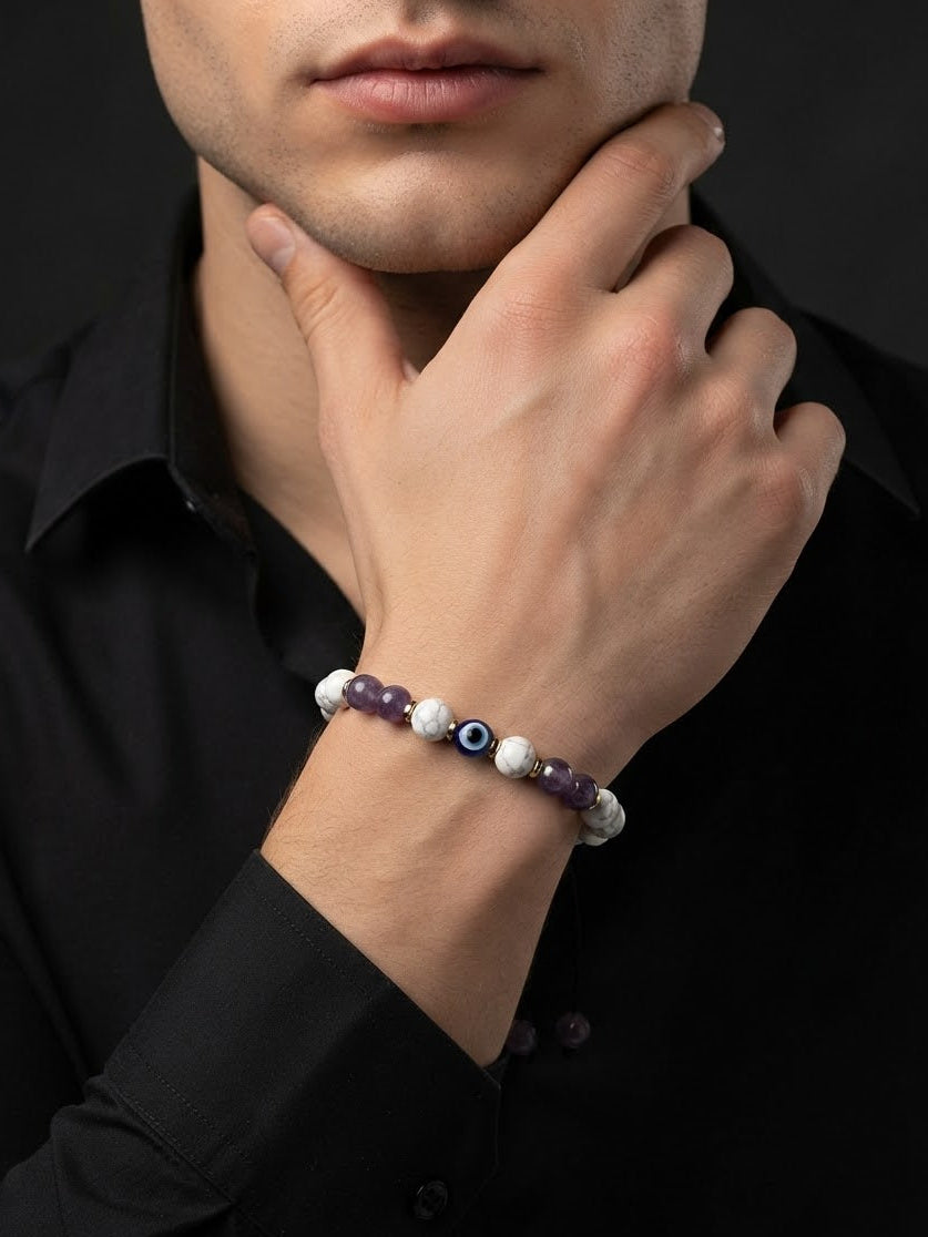 A close-up of a man wearing a beaded bracelet on his wrist, resting his hand on his chin. The bracelet features dark purple beads, white marbled beads, and a central evil eye charm.