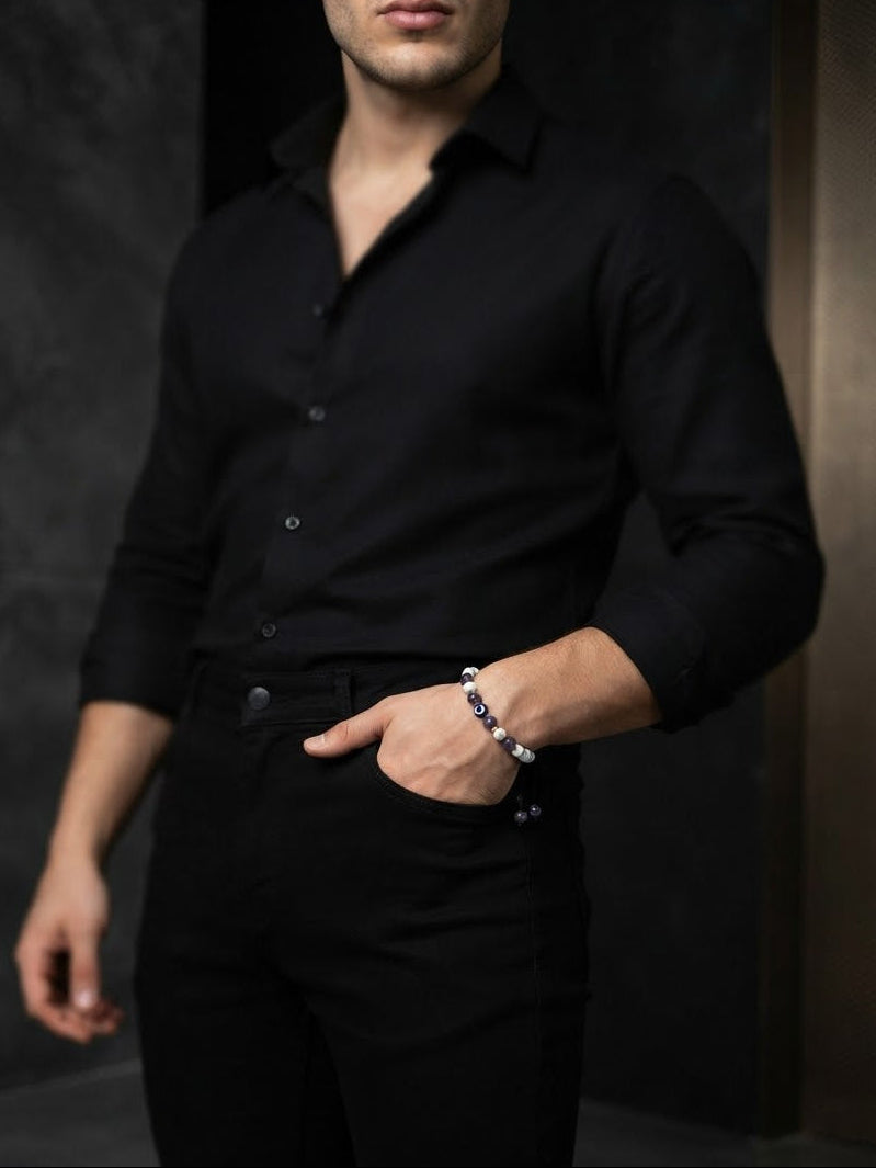 A man wearing a black long-sleeve button-up shirt and black pants, with one hand in his pocket and the other wearing a beaded bracelet, set against a dark textured background.