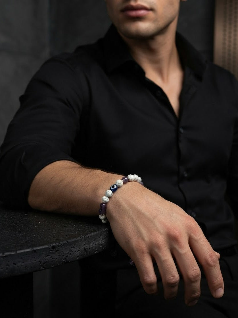 A man wearing a black button-down shirt and a beaded bracelet with white, dark blue, and purple beads, resting his arm on a dark table.