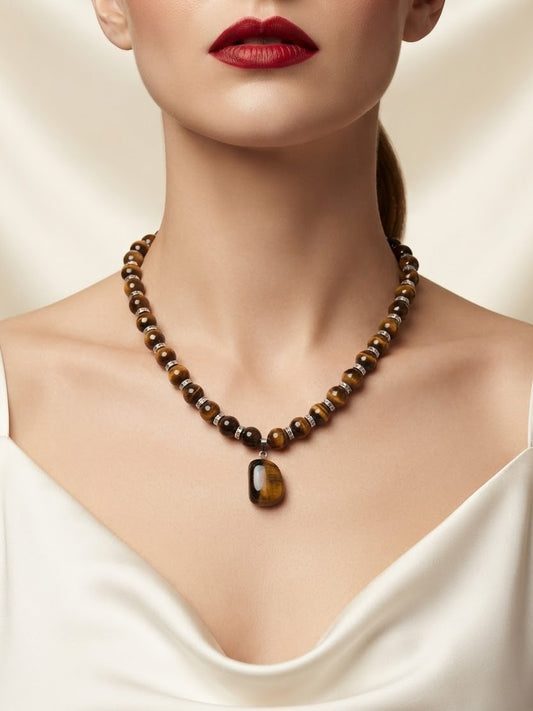 A close-up of a model wearing a beaded necklace with a teardrop-shaped pendant. The necklace features polished brown and black tiger's eye beads with a matching brown pendant.