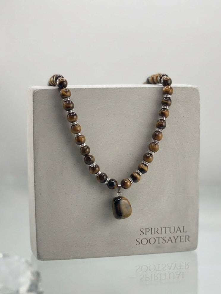 A beaded spiritual necklace with brown and black tiger's eye beads and a polished dark stone pendant, displayed on a square gray stand labeled 'SPIRITUAL SOOTSAYER'.