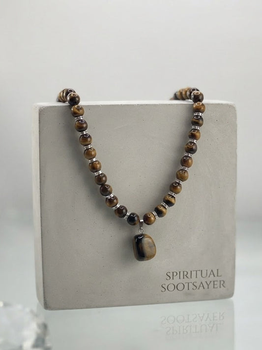 A beaded spiritual necklace with brown and black tiger's eye beads and a polished dark stone pendant, displayed on a square gray stand labeled 'SPIRITUAL SOOTSAYER'.