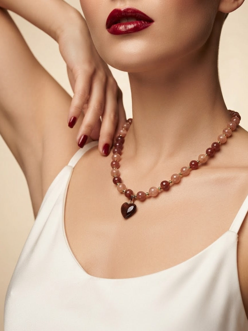 A close-up of a woman wearing a beaded necklace with a dark heart-shaped pendant. The necklace features translucent dusty rose and reddish-brown beads, and the model is wearing a white camisole against a warm beige background.