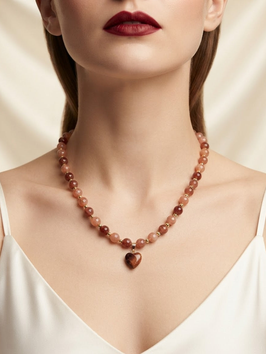 A close-up of a woman wearing a beaded necklace with a heart-shaped pendant. The necklace features warm reddish-brown and peachy-orange beads, and the pendant is a polished dark reddish-brown heart.