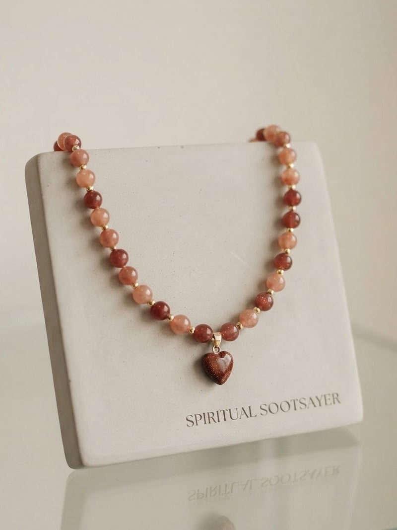A beaded necklace with reddish-brown and peach-orange stones, featuring a small heart-shaped pendant, displayed on a white jewelry stand labeled 'SPIRITUAL SOOTSAVER'.
