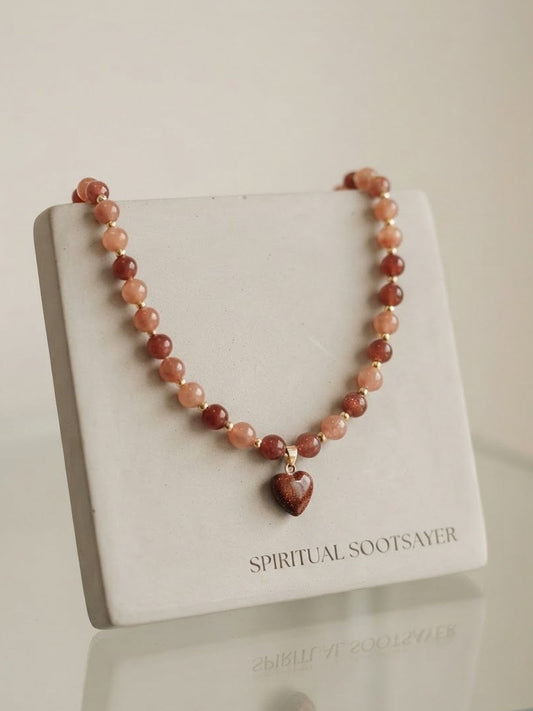 A beaded necklace with reddish-brown and peach-orange stones, featuring a small heart-shaped pendant, displayed on a white jewelry stand labeled 'SPIRITUAL SOOTSAVER'.