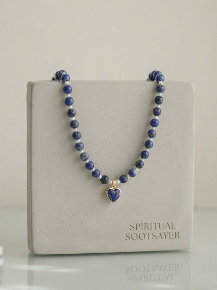 A beaded necklace with dark blue lapis lazuli stones and a small heart-shaped pendant, displayed on a light gray jewelry box labeled 'SPIRITUAL SOOTSAYER'.