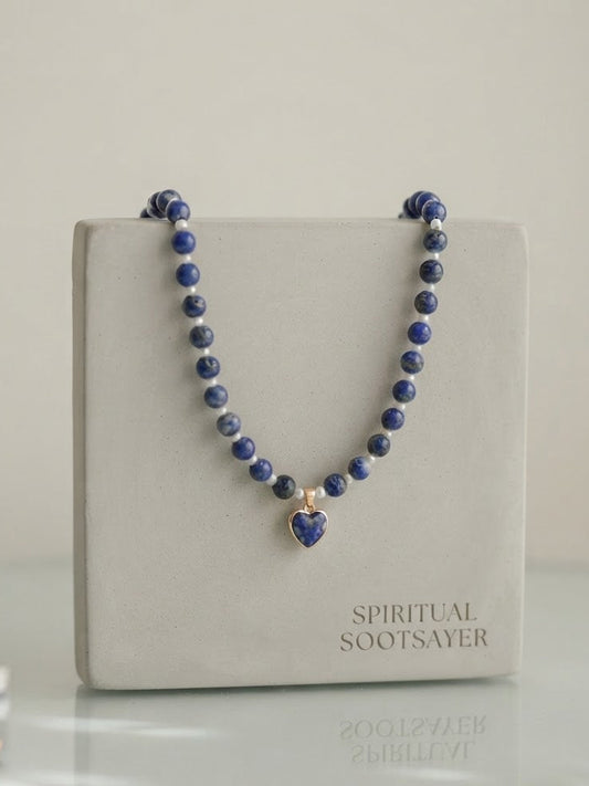 A beaded necklace with dark blue lapis lazuli stones and a small heart-shaped pendant, displayed on a light gray jewelry box labeled 'SPIRITUAL SOOTSAYER'.