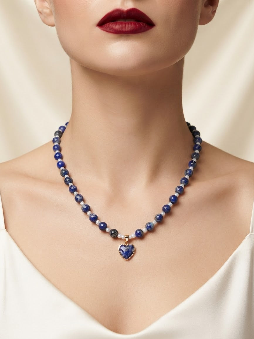 A close-up of a model wearing a beaded necklace with dark blue and white beads and a heart-shaped blue pendant with gold detailing, paired with a white top and red lipstick.